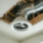 Ceiling vent with silver ductwork exposed through a ceiling opening, surrounded by wooden beams. Neutral tones, industrial setting.