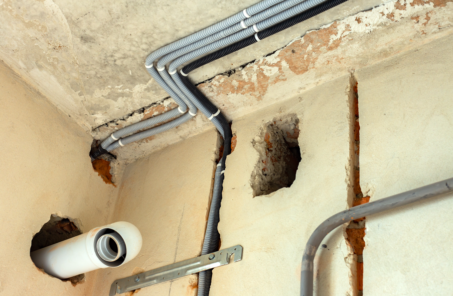 Exposed electrical conduits on a damaged wall with visible bricks. A white pipe juts out from a hole. The mood is industrial and unfinished.