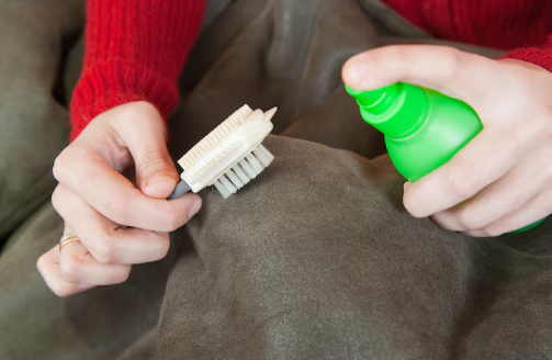 Hands in a red sweater clean suede fabric with a brush and green spray bottle. Cozy setting with emphasis on cleaning action.