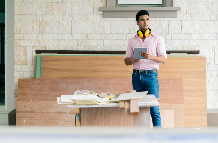 Man in a pink shirt holding a tablet, with yellow headphones around his neck, stands in a workshop with wood panels. Focused expression.