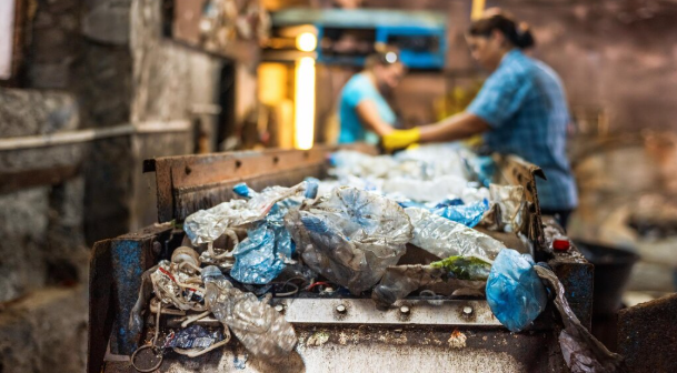 Workers in a recycling facility sort plastic waste on a conveyor. The setting is industrial with blue and yellow accents. Focus on waste.