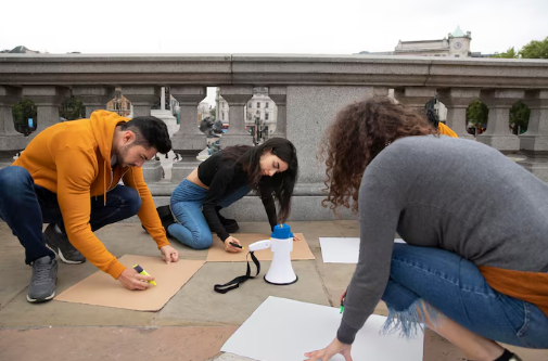 Three people crouch on a stone walkway, creating signs with markers. A megaphone and city buildings are visible in the cloudy background.