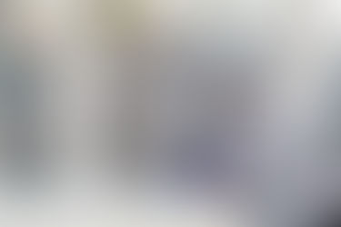 Man wearing a plaid shirt and blue gloves uses a caulking gun on a window frame. Background shows bare trees outside. Calm atmosphere.