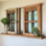Wooden window frames mounted on a wall shelf, with glass vases holding green floral arrangements. Beige wall backdrop. Rustic decor.