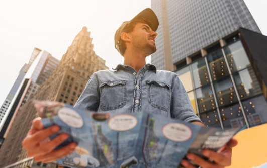 Man in denim shirt and cap holds a map, smiling in a city with skyscrapers in the background. Bright, sunny day. Urban exploration mood.