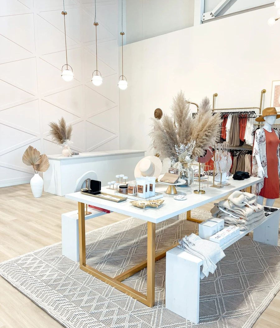 Chic boutique display with decor items on a white table, pampas grass, hanging lights, and clothes. Neutral tones with geometric rug.