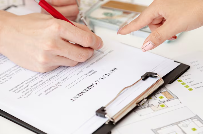Hands sign a rental agreement on a clipboard with a red pen. Background includes floor plans and soft light, conveying a serious tone.