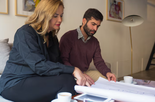 Two people intently review blueprints on a table in a cozy living room. A lamp and paintings decorate the space, evoking a focused mood.