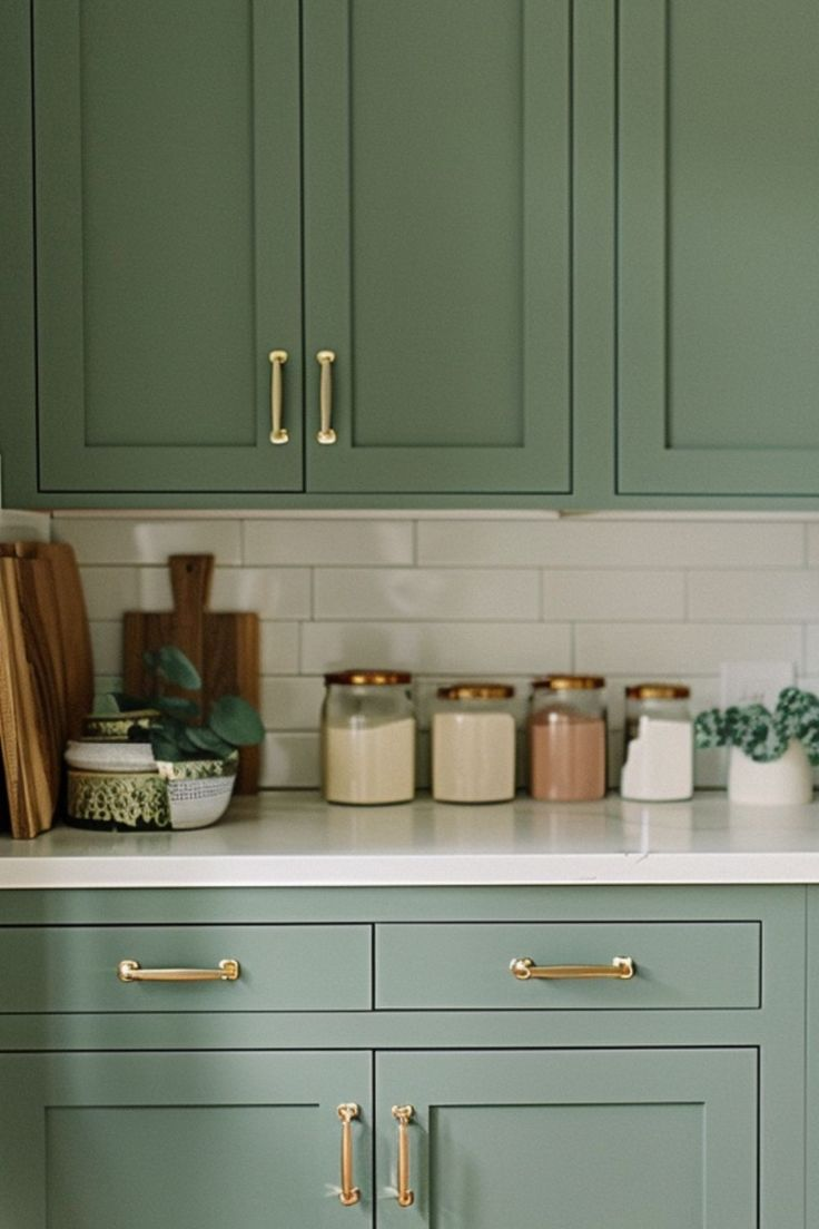 Kitchen with sage green cabinets, gold handles, and a white countertop. A clean white subway tile backsplash is visible, along with neatly arranged jars and cutting boards, creating a minimalist and organized look.
