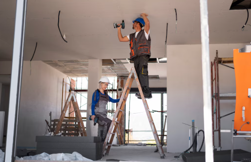 Two construction workers on ladders in a room, one drilling into the ceiling. Both wear safety gear. Scaffolding and tools are visible.