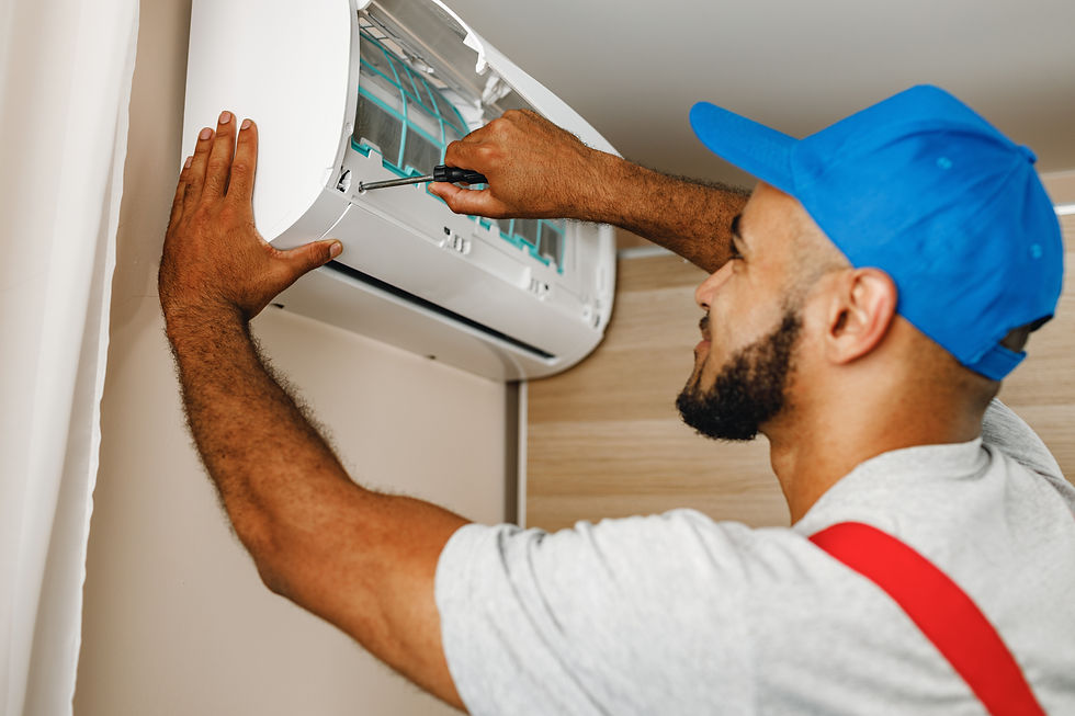Man in blue cap repairs white air conditioner with screwdriver in a beige room. Wears gray shirt and red suspenders, focused expression.