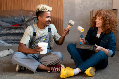 Two people sitting on a floor, holding paintbrushes and rollers, smiling. Background: cardboard boxes and plastic-covered items. Casual mood.