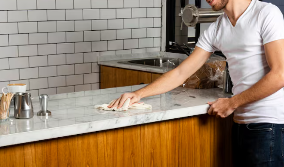 Man cleaning a marble counter with a cloth in a kitchen. White subway tiles in the background. Cup and utensils on the counter. Relaxed mood.