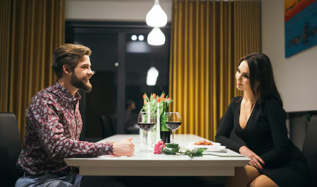 A man and woman sit at a dinner table with wine and flowers. The room has yellow curtains, and a relaxed mood prevails with pendant lights above.