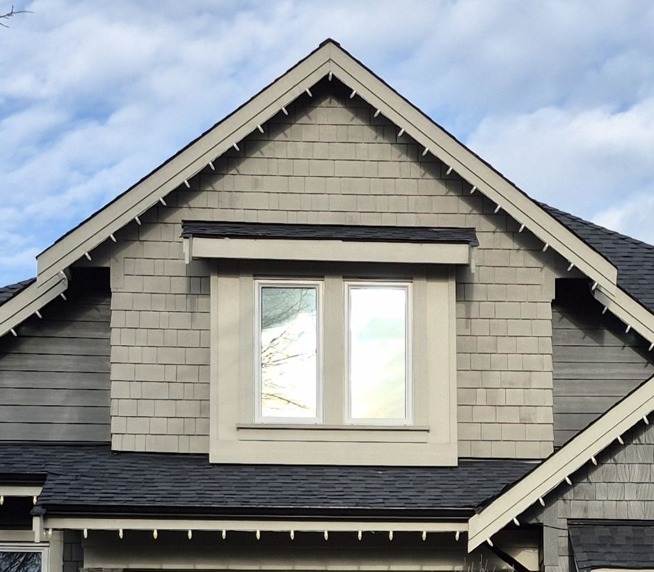 Gray gable house with shingled siding, white-framed window reflecting sky and trees. Overcast sky in the background.