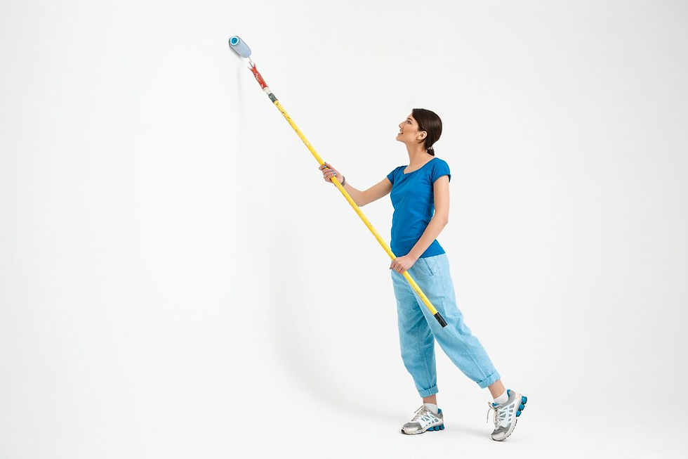 Woman in blue shirt and jeans paints a white wall with a roller on a long handle. She appears focused. Sneakers complete her casual look.