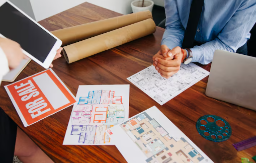 Two people at a table with blueprints, a laptop, a "FOR SALE" sign, and a tablet, discussing plans. Warm lighting; professional setting.