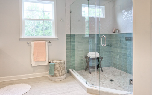 Bright bathroom with a glass shower, teal tiles, and a stool. Towels hang on a rack, and a basket and window add a serene touch.