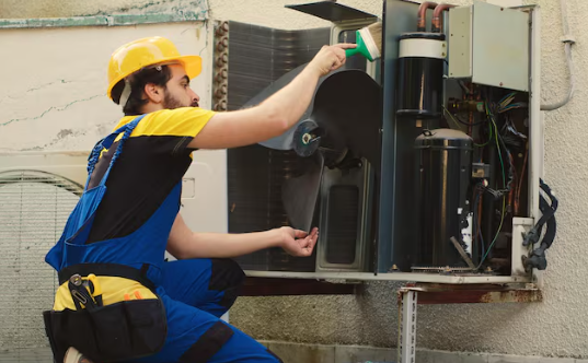 Worker in blue overalls and a yellow hard hat kneels, cleaning an outdoor AC unit with a brush. Tools are visible on his belt.