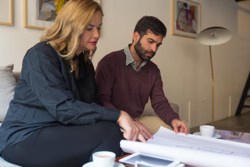 Two people review architectural plans on a table in a modern living room. Calm focus, soft lighting, art on walls, two cups of coffee nearby.