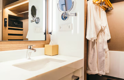 Modern bathroom with a white sink, wall-mounted mirrors, and a beige robe hanging on hooks. Warm lighting creates a cozy ambiance.