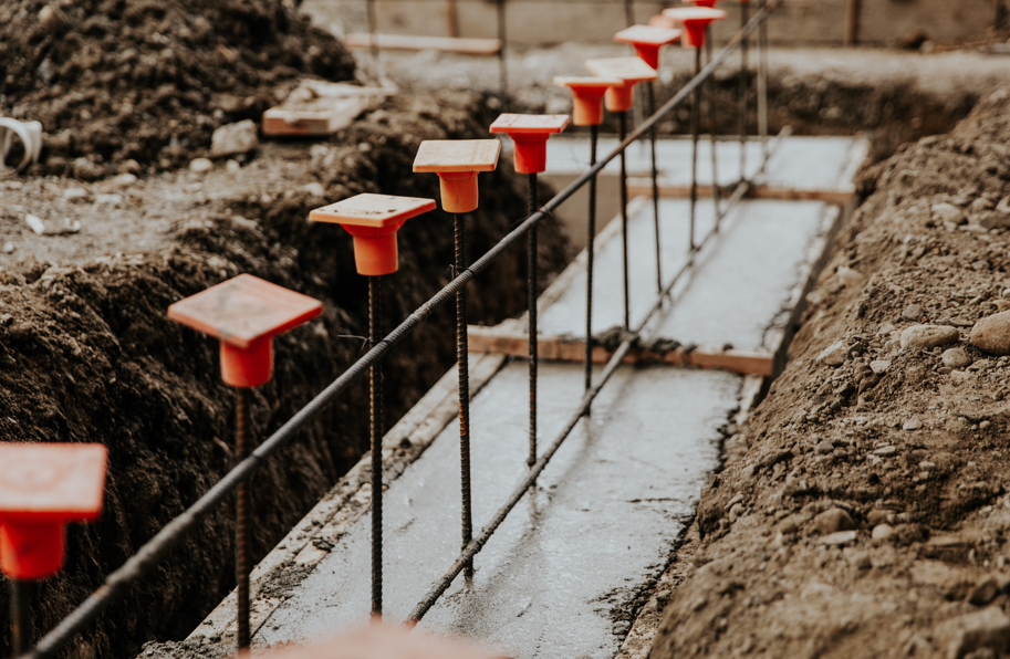 Rebar with orange caps protrudes from a concrete foundation at a construction site. Earth piles line the trench, creating a rugged setting.