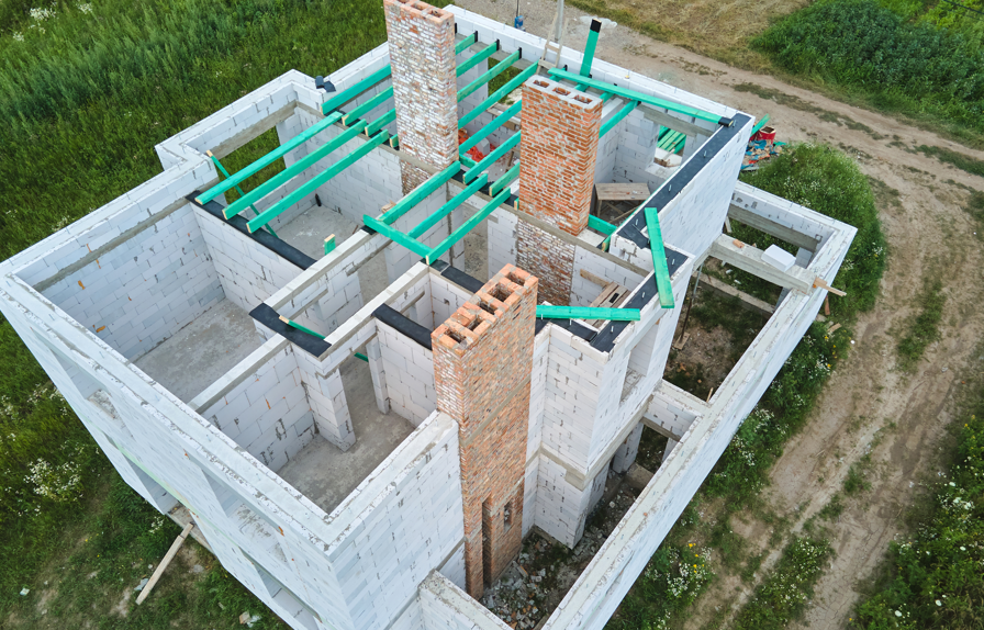 Aerial view of a house under construction with white brick walls and green beams. Brick chimneys visible, surrounded by grass and dirt paths.