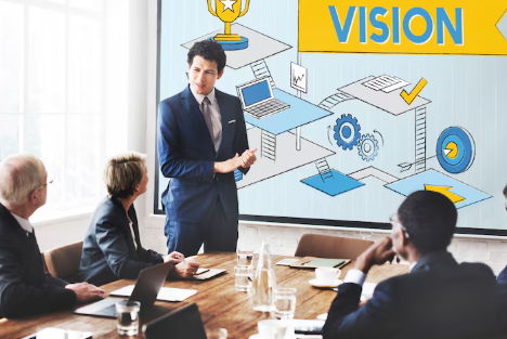 A man in a blue suit presents to colleagues in a conference room. The screen displays "VISION" with gears and icons, fostering focus.