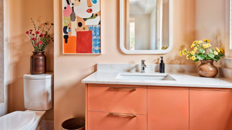 Bathroom with peach cabinets, vibrant abstract art, and flowers in vases. A large mirror reflects light, creating a fresh, colorful mood.