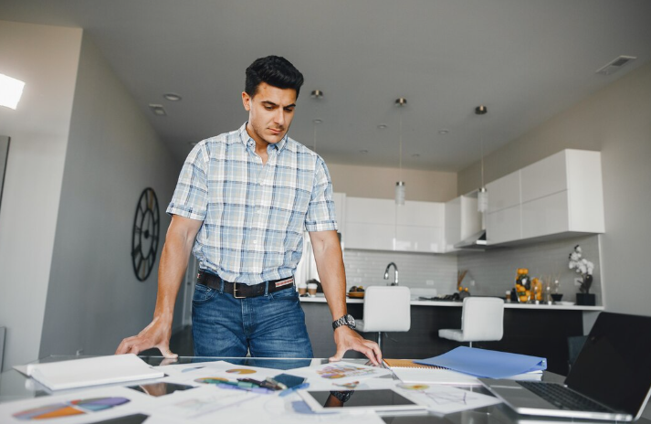 Man in plaid shirt studies papers on a desk in a modern kitchen. Laptop open, atmosphere focused, neutral tones, and minimalistic decor.