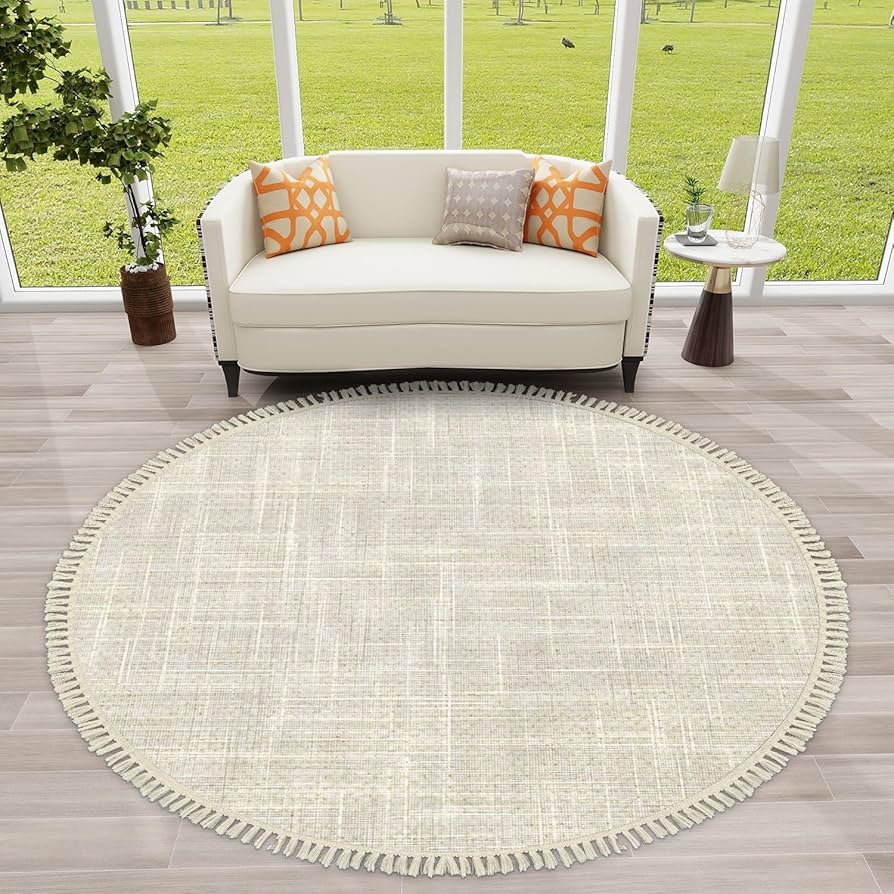 A round beige rug on wooden floor, white sofa with orange cushions, and a side table with a lamp and plant. Large windows show green lawn.