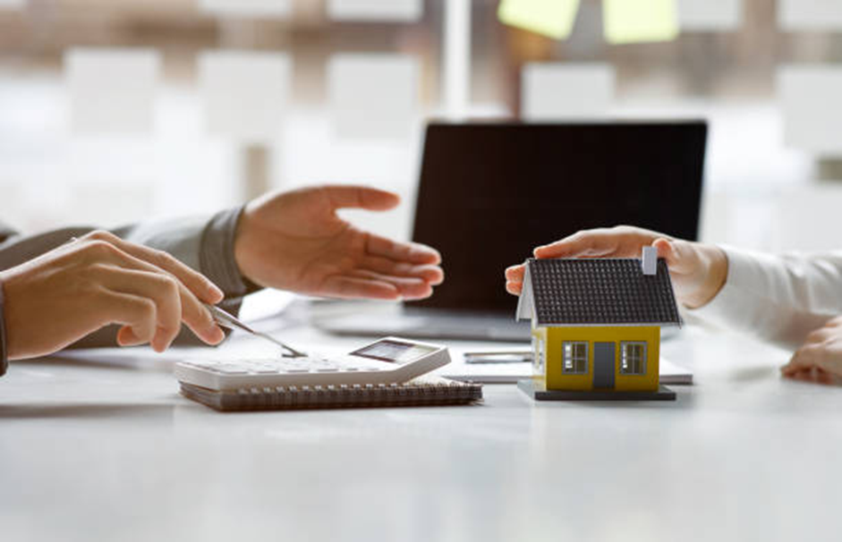 Hands discuss over papers and a calculator on a desk. One hand holds a small yellow house model. Laptop in the background, office setting.
