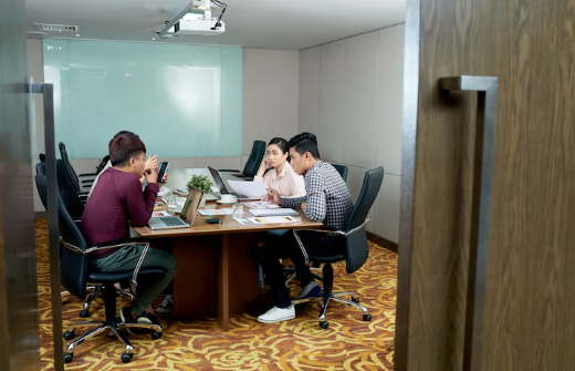 Four people in a conference room, sitting at a table with laptops and documents. The setting has patterned carpet and neutral walls.