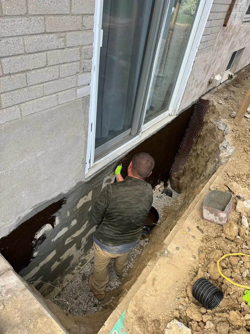 A man in a camo jacket applies sealant to a house foundation in a trench. The setting shows exposed dirt, gravel, and construction tools.