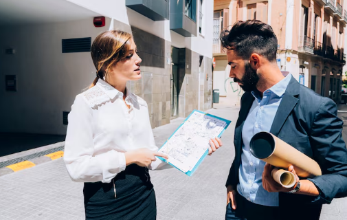 A woman and man in business attire discuss a clipboard map on a sunlit street. He holds rolled-up papers. Buildings in the background.