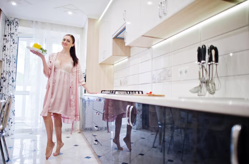 Woman in pink robe holds fruit on a plate, smiling in a modern kitchen. White cabinets, knife set, and sunny window in the background.