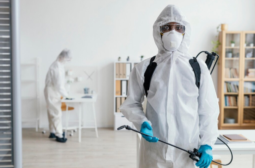 Two people in hazmat suits clean a light-colored room. One holds a sprayer; the other works at a desk. Background shows shelves and books.