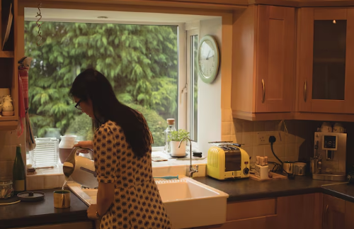 Woman in a patterned dress pours tea in a cozy kitchen with wooden cabinets. A window shows greenery outside. A clock and a yellow toaster are visible.