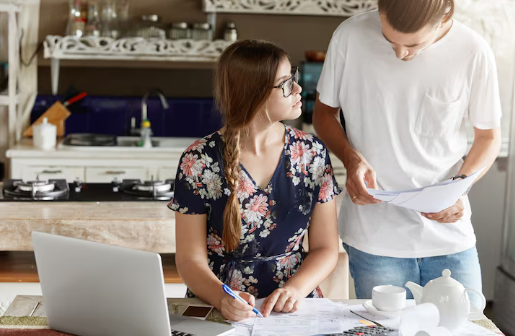 Woman in floral dress writes at table with laptop and papers. Man in white shirt looks at documents. Kitchen setting, casual mood.