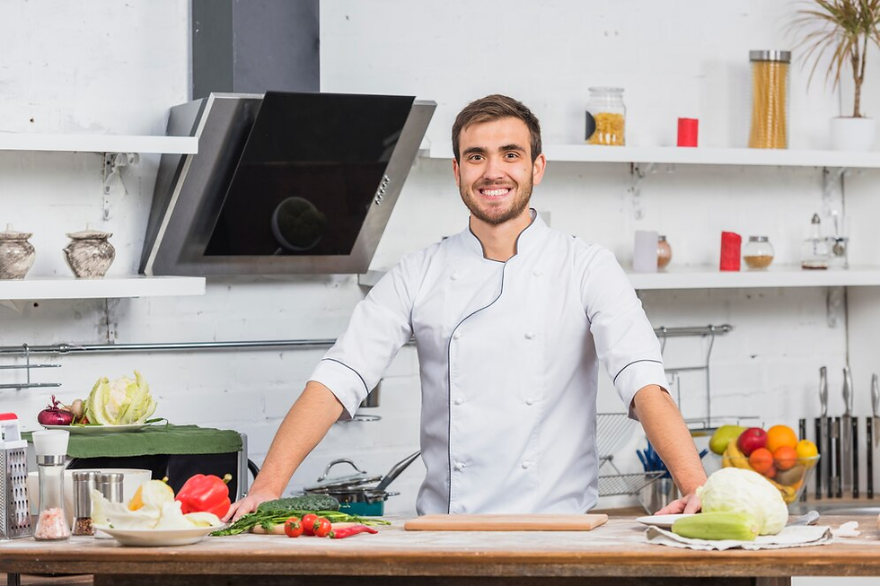 Chef in white uniform smiles in a kitchen. Vegetables and fruits on counter, shelves with jars in background. Bright, cheerful atmosphere.