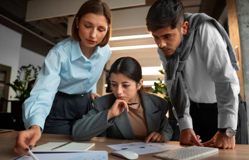 Three people in an office analyze documents on a table. Two stand and one sits, focused and engaged. Background features plants and lighting.