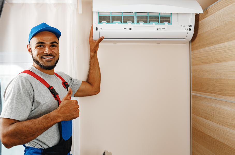 Smiling technician in blue cap gives a thumbs-up beside an air conditioner mounted on a pale wall, wearing grey shirt and red straps.