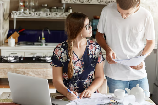 Two people reviewing documents in a kitchen. Woman in floral dress, man in white shirt. Open laptop, teapot on table. Focused mood.