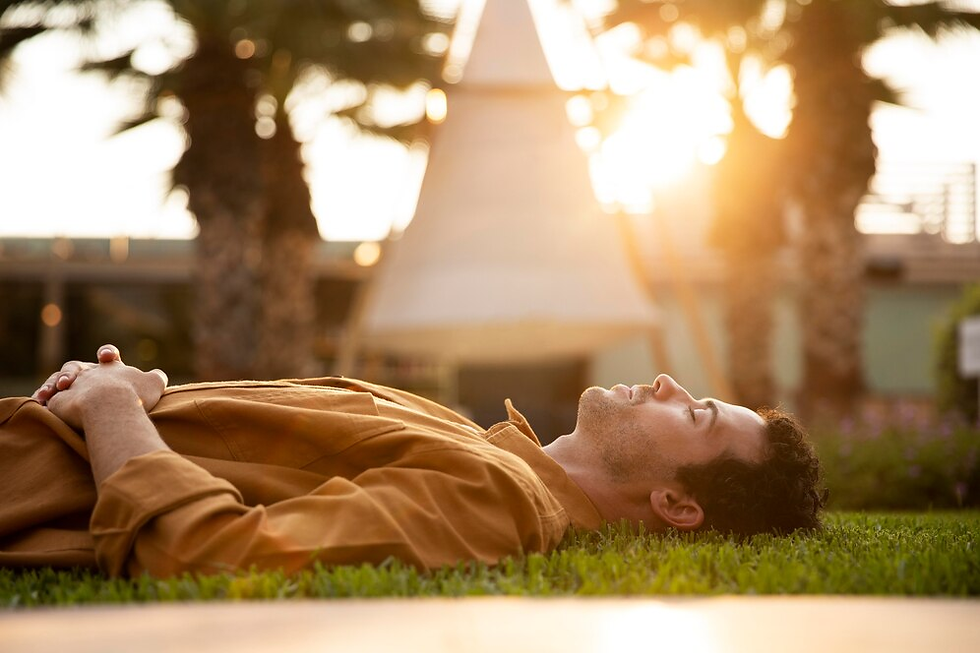 Man in orange shirt lying on grass, eyes closed, basking in warm sunlight. Palm trees and sunlight in the background create a serene vibe.