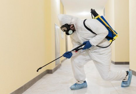 Person in protective gear sprays disinfectant in a hallway. Wearing white suit, gas mask, blue gloves, boots. Yellow walls, focused action.