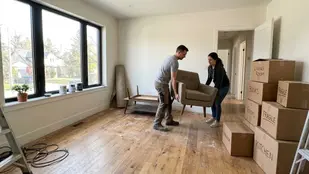 A man and woman move an armchair in a bright room with wooden floors. Labeled boxes stack nearby. Large window and unpacked items visible.