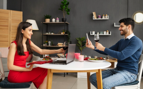 Couple having a virtual dinner date with wine, smiling at each other through a laptop. Cozy room with candles and plants in the background.