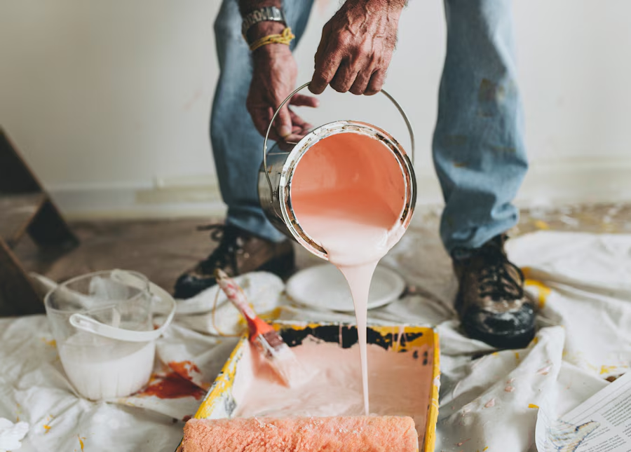 Person pouring pink paint into a tray on a drop cloth. Paint roller and other tools visible. Casual setting with jeans and boots.