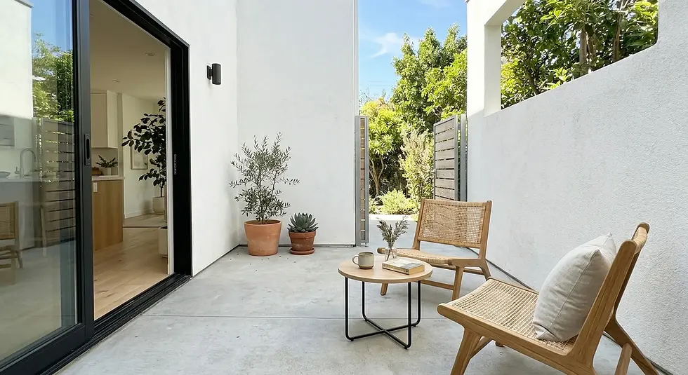 Outdoor patio with two wicker chairs, a small table holding a mug and book, potted plants, white walls, and open sliding glass door. Bright, serene setting.