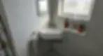 Small bathroom with white corner sink, towel, toothbrushes, soap dispenser, and a potted plant on the windowsill. Gray tiles and soft light.